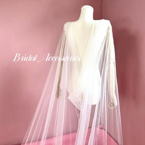 Pu&ograve; includere: Un mantello da sposa in tulle bianco con schiena bassa e un delicato motivo a pois. Il mantello &egrave; drappeggiato su un manichino ed &egrave; mostrato su uno sfondo rosa.
