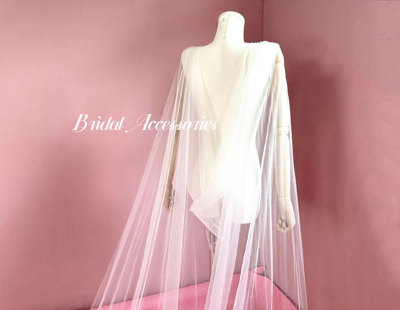 Pu&ograve; includere: Un mantello da sposa in tulle bianco con schiena bassa e un delicato motivo a pois. Il mantello &egrave; drappeggiato su un manichino ed &egrave; mostrato su uno sfondo rosa.