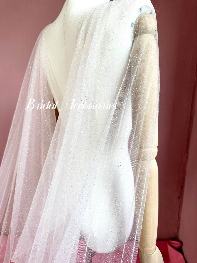 Pu&ograve; includere: Velo da sposa in tulle bianco con punti di glitter argentati. Il velo &egrave; drappeggiato su un manichino.