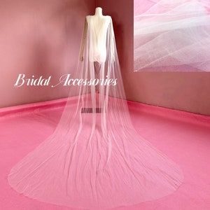 Pu&ograve; includere: Un mantello da sposa in tulle bianco con un tessuto trasparente e scintillante. Il mantello &egrave; drappeggiato su un manichino e ha una lunga coda.