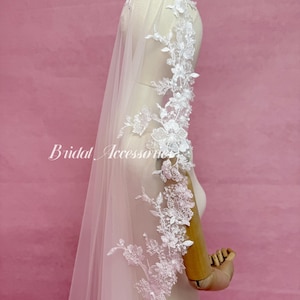 May include: A pale pink bridal veil featuring intricate white floral lace embroidery along the edges, accented with small clear beads and sequins. The veil is draped over a mannequin, and the text Bridal Accessories is visible on the left side.