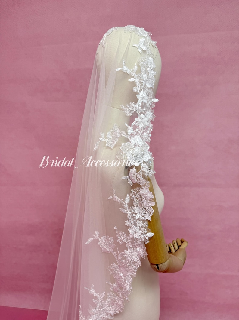 May include: A pale pink bridal veil featuring intricate white floral lace embroidery along the edges, accented with small clear beads and sequins. The veil is draped over a mannequin, and the text Bridal Accessories is visible on the left side.