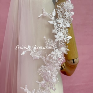 May include: A white bridal veil featuring intricate floral lace embroidery, clear sequins, and small beads along the edge. The veil is draped over a mannequin, and the text Bridal Accessories is printed in white script across the center of the fabric.