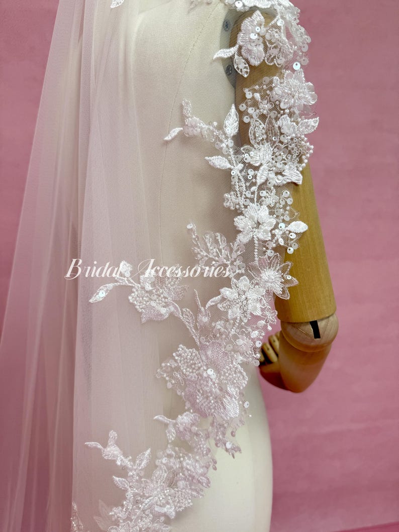May include: A white bridal veil featuring intricate floral lace embroidery, clear sequins, and small beads along the edge. The veil is draped over a mannequin, and the text Bridal Accessories is printed in white script across the center of the fabric.