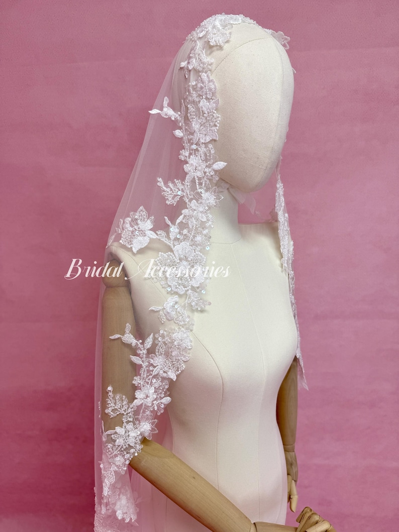 May include: A white bridal veil featuring intricate floral lace embroidery along the edges, accented with small sequins and beads. The veil is draped over a mannequin, and the text Bridal Accessories is printed in white on the left side.