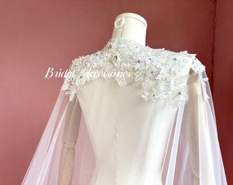 May include: A white bridal cape with a delicate lace overlay and beaded embellishments. The cape is made of sheer tulle and has a flowing silhouette.