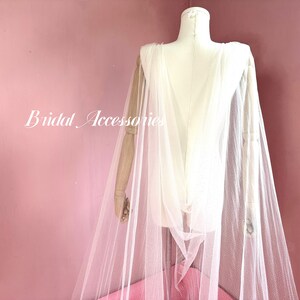 Pu&ograve; includere: Un mantello da sposa in tulle bianco con un delicato motivo a pois. Il mantello &egrave; drappeggiato su un manichino e ha un design lungo e fluido.