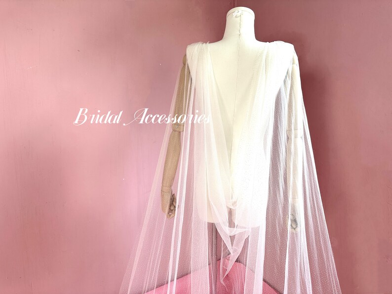 Pu&ograve; includere: Un mantello da sposa in tulle bianco con un delicato motivo a pois. Il mantello &egrave; drappeggiato su un manichino e ha un design lungo e fluido.