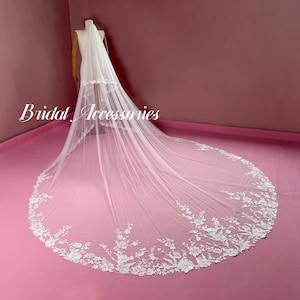 May include: A long, white bridal veil with a floral lace border is displayed on a mannequin. The veil is sheer and delicate, with a train that extends across a pink surface. The words "Bridal Accessories" are written in cursive.