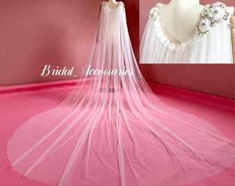 May include: A white tulle bridal cape with a beaded embellishment on the back. The cape is draped over a mannequin and is laying on a pink surface. The text "Bridal Accessories" is visible in the image.