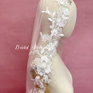 May include: A white bridal veil featuring intricate floral lace embroidery along the edges. The lace is embellished with small clear sequins and beads. The veil is draped over a mannequin head and shoulders. The text Bridal Accessories is visible.