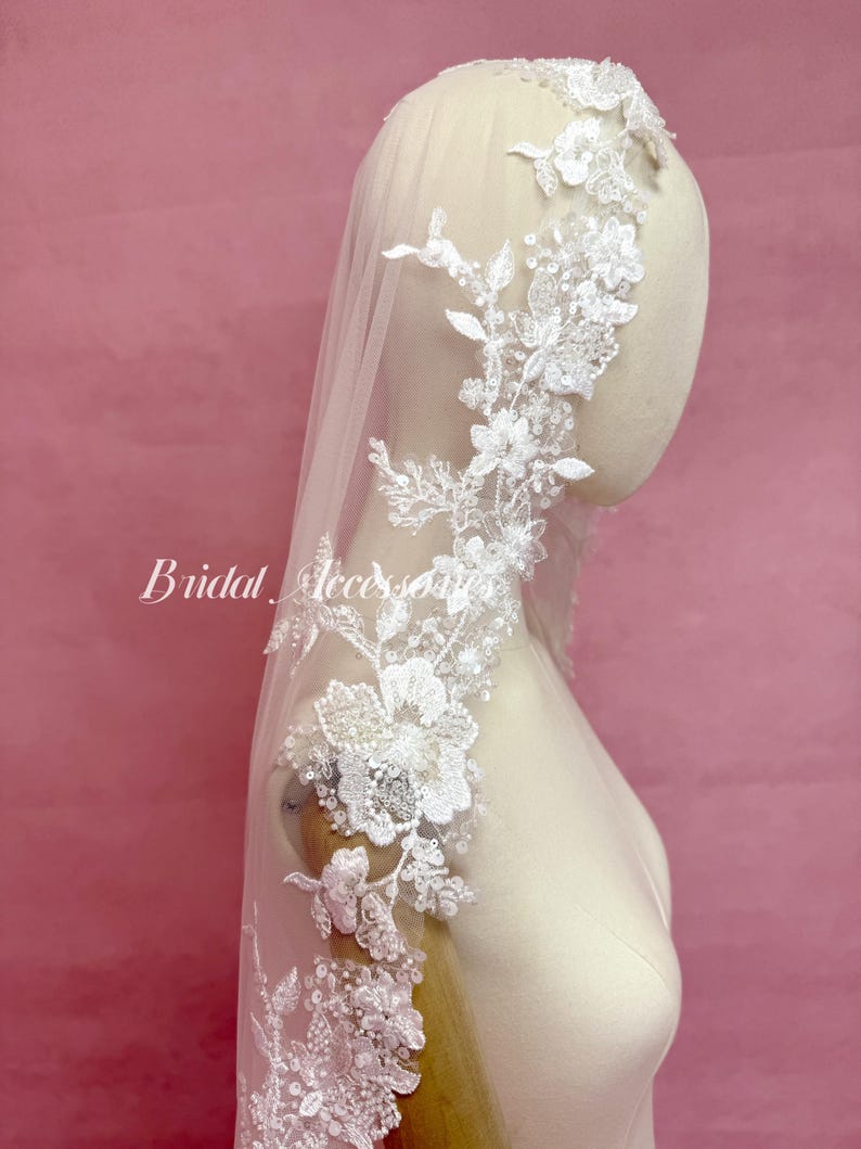 May include: A white bridal veil featuring intricate floral lace embroidery along the edges. The lace is embellished with small clear sequins and beads. The veil is draped over a mannequin head and shoulders. The text Bridal Accessories is visible.