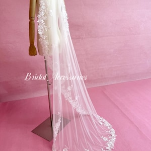 May include: A long white tulle wedding veil featuring intricate floral lace embroidery along the edges. The veil is draped over a mannequin and displays the text Bridal Accessories in white script. The delicate lace pattern extends from the top.