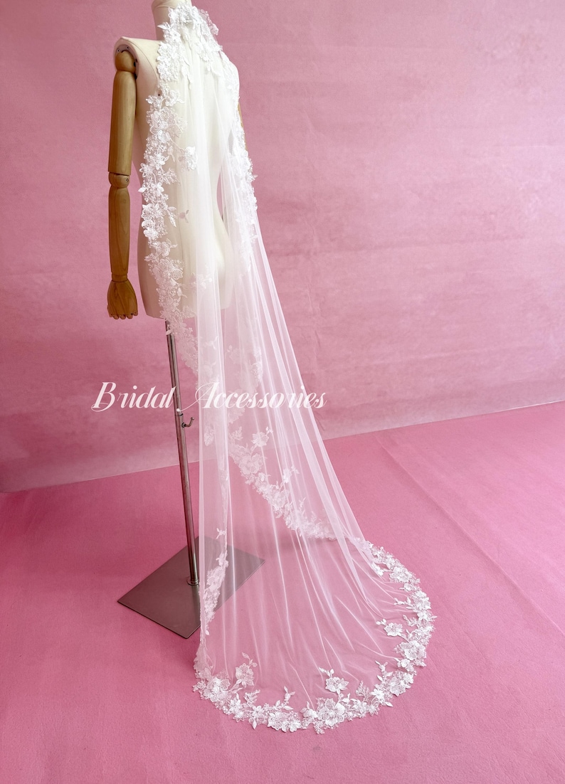 May include: A long white tulle wedding veil featuring intricate floral lace embroidery along the edges. The veil is draped over a mannequin and displays the text Bridal Accessories in white script. The delicate lace pattern extends from the top.