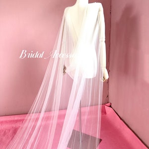 Pu&ograve; includere: Un mantello da sposa in tulle bianco con una lunga coda, drappeggiato su un manichino bianco su sfondo rosa. Il mantello &egrave; trasparente e delicato, con una texture morbida e fluida.