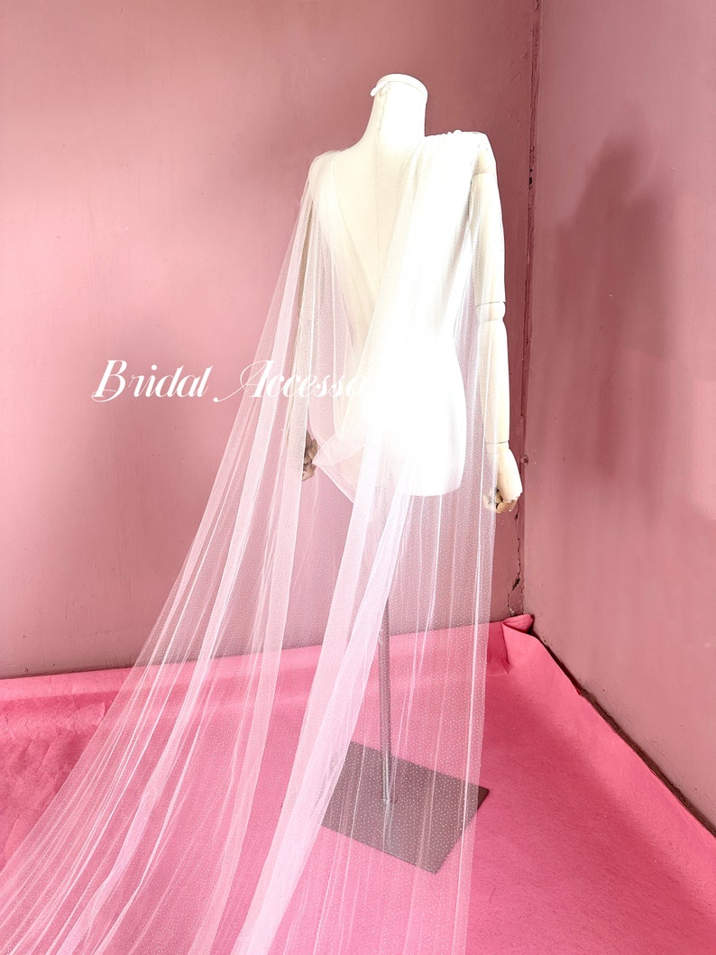 Pu&ograve; includere: Un mantello da sposa in tulle bianco con una lunga coda, drappeggiato su un manichino bianco su sfondo rosa. Il mantello &egrave; trasparente e delicato, con una texture morbida e fluida.