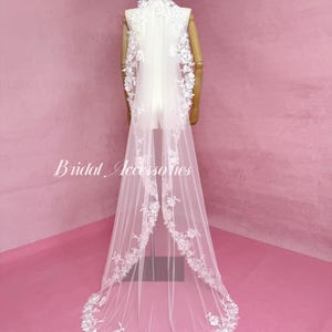 May include: A long white bridal veil featuring intricate floral lace embroidery along the edges and a high collar neckline. The sheer tulle fabric drapes down to the floor, with the text Bridal Accessories visible on the left side of the image.