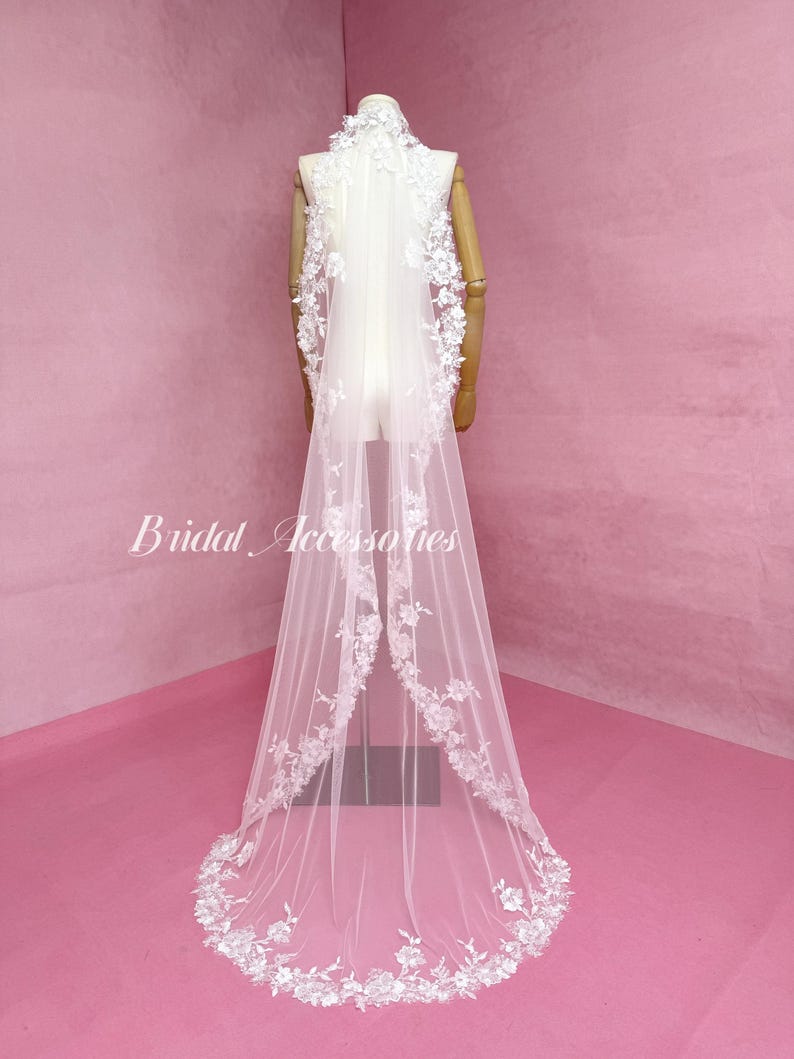 May include: A long white bridal veil featuring intricate floral lace embroidery along the edges and a high collar neckline. The sheer tulle fabric drapes down to the floor, with the text Bridal Accessories visible on the left side of the image.