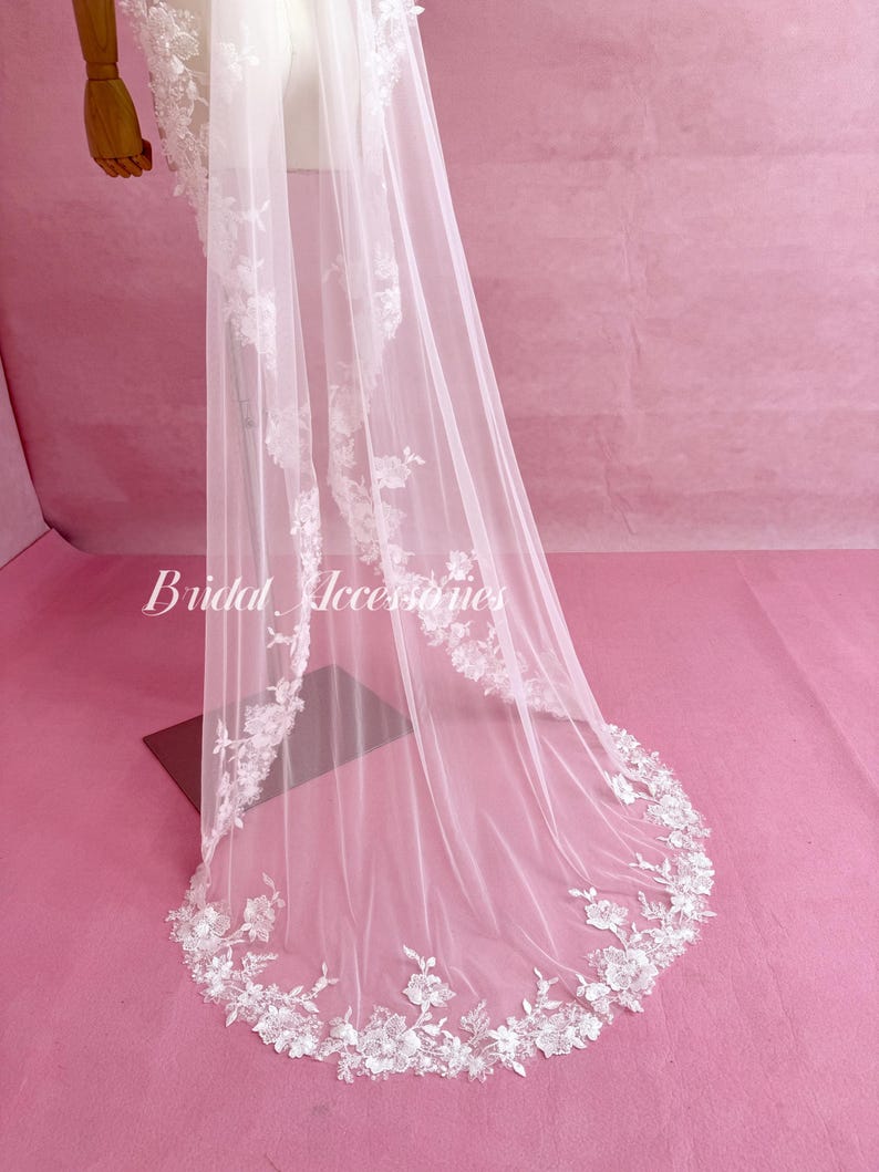 May include: A long, sheer white veil featuring intricate white floral lace embroidery along the edges. The delicate lace pattern extends from the top down to the rounded hem. The text Bridal Accessories is printed in white on the left side.
