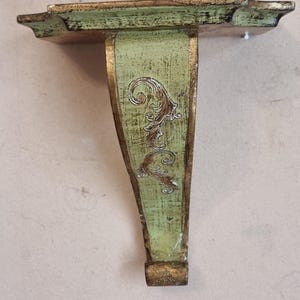 Peut inclure: Une étagère murale décorative avec une finition verte vieillie et des accents dorés. L'étagère a un dessus rectangulaire et un seul support orné. L'étagère mesure environ 25 cm de large.