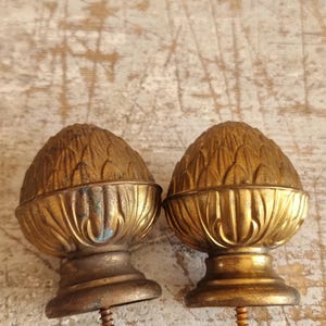 Op de afbeelding: Twee antieke goudkleurige finials, elk met een gedetailleerd, gestructureerd ontwerp dat lijkt op een dennenappel. De finials hebben een decoratieve basis en een schroef voor bevestiging. De objecten staan op een verweerd houten oppervlak.
