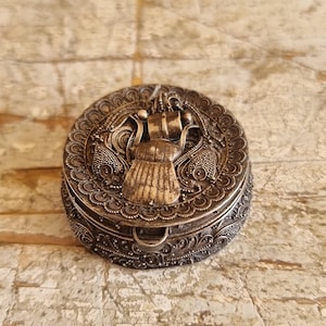 May include: An ornate silver-coloured round trinket box with a detailed lid featuring a central relief design. The box has a decorative border and a small clasp. The background is a textured, light-coloured surface.