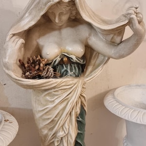 Op de afbeelding: Klassiek beeld van een vrouw met een gedrapeerde sluier en een gouden kroon. Het beeld houdt een mand met bloemen en kralen vast. De figuur is geschilderd in crème-, groen- en goudtinten.