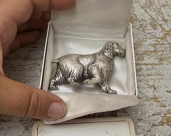May include: A silver-toned metal dog figurine displayed inside a white satin-lined box. The box lid features the gold-colored text J.D.F. Figurines, 8 Rue Borie, Bordeaux. The dog is depicted in a standing position with detailed fur texture.