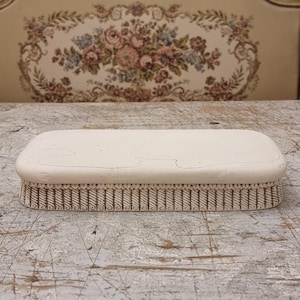 May include: An off-white rectangular brush with a patterned border, set on a distressed wooden surface. The brush's design suggests a vintage or antique style. The surface is light-colored and shows signs of wear.