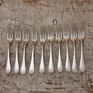 May include: Ten silver forks displayed in a row. Each fork features a slender handle with a decorative, fan-shaped design at the base. The forks are set against a weathered, grey wooden surface.