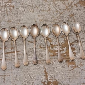 Peut inclure: Ensemble de neuf cuillères en argent disposées sur une surface en bois vieilli. Les cuillères ont des cuillères ovales et des détails décoratifs en forme de coquillage sur les manches. Elles présentent des signes d'usure.