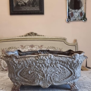 Puede incluir: Una jardinera rectangular y desgastada con relieve floral ornamentado. El recipiente gris, apoyado sobre cuatro pequeños pies, tiene un borde decorativo. Probablemente de hierro fundido, es apta para uso interior o exterior. Medidas en centímetros.