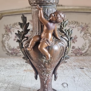 Puede incluir: Un jarrón de metal en tonos bronce que presenta la figura central de un querubín alcanzando un borde de cestería adornado con uvas y hojas esculpidas. El cuerpo del jarrón muestra detalles en relieve florales y de follaje sobre una base.