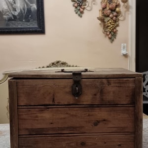 May include: A rectangular wooden storage box constructed from horizontal planks with a visible wood grain. The box features a hinged lid secured by a dark, weathered metal latch centered on the front panel. The wood has a natural brown finish.