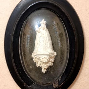May include: A framed religious relief sculpture featuring a white figure holding a child, set against a glass background. The black oval frame has a metal loop for hanging. The sculpture is likely a depiction of a religious figure.