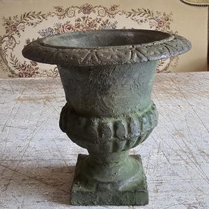 May include: A weathered green urn with a wide, flared rim featuring a repeating geometric pattern. The body of the urn has a textured, mottled finish with a bulbous midsection and a square base, standing as a decorative vessel for plants.