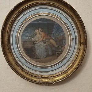 Peut inclure: Un cadre circulaire doré et orné contenant une estampe de style ancien représentant deux personnages en vêtements d'époque s'embrassant près d'un piano. L'image présente des tons sourds de bleu, brun et crème, avec du texte visible en bas.