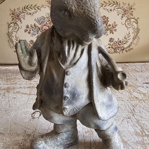 May include: A gray, weathered-finish rabbit figurine standing upright. The rabbit is dressed in a buttoned waistcoat and jacket, with one hand raised in a gesture and the other holding a pipe. It features textured fur details and a vintage appearance.