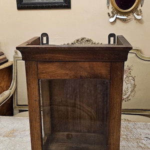 Peut inclure: Une vitrine murale rectangulaire en bois dotée d'une façade en verre transparent. Le cadre est fabriqué en bois de ton moyen avec des motifs de grain visibles. Deux supports de fixation en métal noir sont fixés sur le bord arrière supérieur.