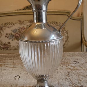 May include: A silver-plated metal and clear glass claret jug featuring a vertical ribbed glass body, a polished silver-tone metal base, a matching silver-tone metal neck with a curved handle, and a pointed spout topped with a finial lid.