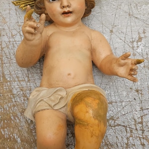 Op de afbeelding: Een geschilderd beeldje van een kind met krullend bruin haar, gekleed in een crèmekleurige lendendoek. Het figuur heeft een goudkleurige stralenkrans achter het hoofd, met één hand opgeheven en de andere hand opzij gestrekt.