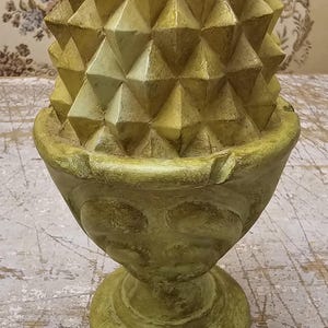 Puede incluir: Un objeto decorativo con forma de piña con una superficie geométrica texturizada en tonos verde y amarillo apagados. La piña descansa sobre una base a juego que presenta un cuenco redondeado y un pie circular con un acabado desgastado.