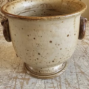 Puede incluir: Una maceta de cerámica grande y redonda con un borde ancho y acampanado y dos asas laterales curvas de color marrón oscuro. El exterior presenta un esmalte blanquecino moteado con manchas marrones visibles y un acabado texturizado y desgastado.