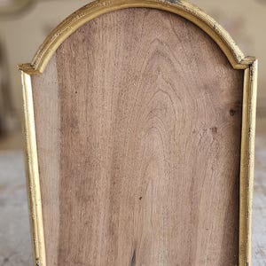 May include: An arched picture frame featuring a distressed gold-toned metal border with intricate detailing. The center panel consists of a flat, light brown wooden surface with a visible natural grain pattern, framed by the metallic gold structure.