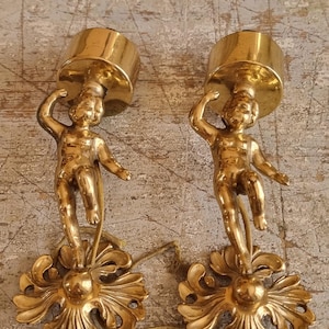 Puede incluir: Dos candelabros antiguos de color dorado. Cada uno presenta una figura de querubín que sostiene una plataforma circular. Los querubines se alzan sobre bases ornamentadas en forma de flor. Los candelabros son de metal y tienen un acabado brillante.