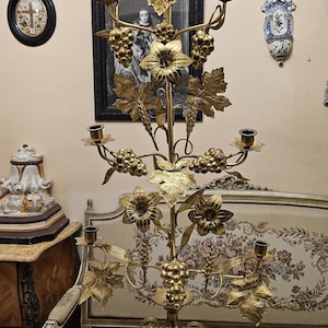 Può includere: Un alto candelabro in ottone decorato con cinque portacandele disposti su una struttura a livelli. Lo stelo centrale è ornato da grappoli d'uva, foglie di vite e fiori in metallo, sostenuto da un'ampia base decorativa.