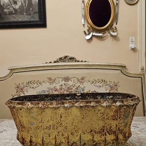 Pode incluir: Uma jardineira oval de metal amarelo com um acabamento bastante desgastado e enferrujado. O recipiente apresenta uma borda superior recortada, detalhes nervurados verticais nas laterais e uma base larga e plana, com sinais de desgaste.