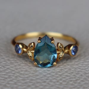 Op de afbeelding: Een gouden ring met een peervormige, lichtblauwe edelsteen als middelpunt. De ring heeft twee kleinere blauwe edelstenen aan weerszijden, met decoratieve bladachtige accenten. De ring is geplaatst tegen een neutrale grijze achtergrond.