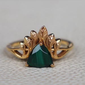 May include: A gold ring featuring a triangular, deep green gemstone. The ring's design includes a decorative leaf-like pattern around the gemstone. The band is a simple, thin gold band.