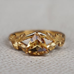 Könnte beinhalten: Ein goldener Ring mit einem dreieckigen, hellorangen Edelstein. Der Ring hat ein florales Design am Band, mit Blättern und filigranen Details. Der Edelstein ist der Mittelpunkt, gefasst in einem goldenen Band.